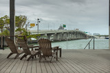 Fototapeta Boho - Sillas en una terraza de madera con vista al mar y al puente con cielo nublado