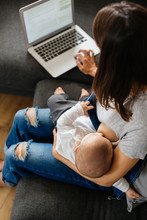 Young Mother Breast Feeding Her Baby Whilst Working On Laptop At Home.