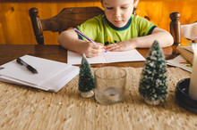 Little Boy Sitting At The Kitchen Table Signing Christmas Cards