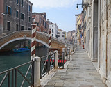 Fototapeta Na drzwi - Venice, Italy, medieval stone street along a canal with ancient buildings, a bridge and the typical white and red poles of the gondola docking