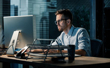 Young Focused Man Doing Overtime Job Watching Computer At Desktop In Dark Office. 