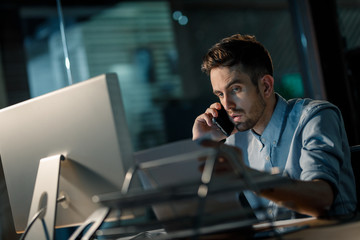 Wall Mural - Formal manager speaking on smartphone working with papers in office late at night sitting at desktop. 