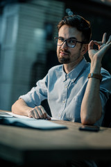 Wall Mural - Stylish young man in glasses sitting at working table in office showing OK gesture and looking confidently at camera