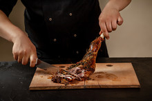 Chef Is Cutting Roasted Leg Lamb On Wooden Board On Table