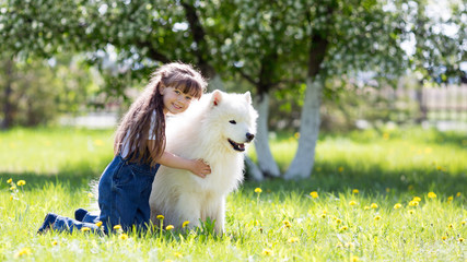 Little girl with a big white dog in the park. A beautiful 5 year old girl in jeans hugs her favorite dog during a summer walk.