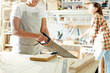 © pressmaster - Crop female woodworker using saw to cut wooden plank while working in joinery.