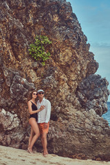 Wall Mural - Attractive couple posing on a beach near big reef stones, enjoys a vacation on a beautiful island. 