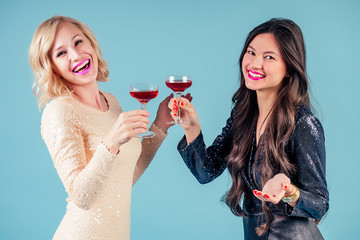 two happy and attractive girlfriends brunette and blonde womens in evening cocktail shiny sequins dress are holding a glass of wine and gossip talk at a party in the studio on a blue background