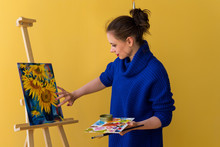 Girl Artist Paints Sunflowers Oil Paints On Canvas. She Is Wearing Blue Sweater. Woman Is Holding Brush And Palette With Paints. She Puts Paint On Her Fingers.