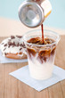 © demphoto - Close-up shot of delicious fresh coffee filling plastic cup with cold milk and ice cream on table in donut cafe