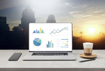 Poster - laptop showing blank screen with cup of coffee and mobile phone on table against beautiful business modern office buildings background. technology concept 