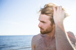 Attractive shirtless man touching hair and looking away while standing on beach near sea.