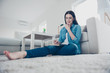 © deagreez - Low angle view of thoughtful pensive woman sitting on carpet near sofa in modern white apartment holding hand on chin writing list to do in notepad using pen wearing jeans outfit