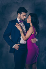 Poster - Portrait of bearded man in tuxedo with bow holding hand in pocket of pants and charming pretty lady with booty ass jewelry, attractive spouses hugging looking at each other over grey background