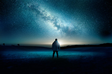 a man stands watching with wonder and amazement as the moon and milky way galaxy fill the night sky.