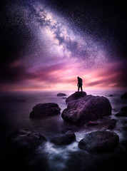 Wall Mural - night time sea landscape with the milky way. a man stands on a rocky shore line with the stars above