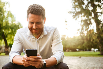 image of handsome guy in white shirt, sitting in park and looking at black mobile phone holding in h