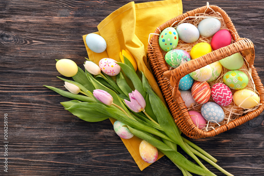 Easter basket with colorful eggs and spring flowers on wooden background, top view
