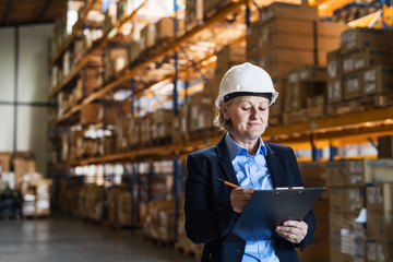 Senior woman warehouse manager or supervisor with white helmet and clipboard.