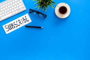 Poster - Email subscribe concept. Hand lettering subcribe on work desk with plant, glasses, computer keyboard, cup of coffee on blue background top view copy space
