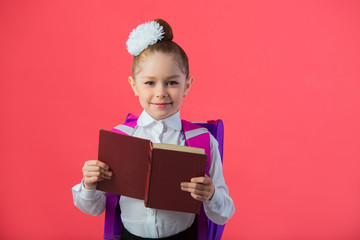 Wall Mural - beautiful little girl with a bow with a school satchel on a pink background with a book in hands