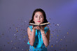 Young woman or teen girl is blowing glitter confetti.Asian girl teenager holds book in hands,on purple background festive sparkles for the party are flying.Concept Christmas,new year,holiday,birthday.