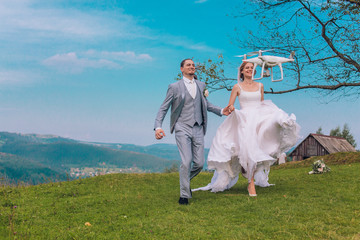 Young coulple running to flying drone. Concept of aerial shooting. White drone flying aroud bride and groom. Advertising picture.