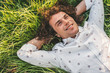 © iuricazac - Above shot portrait of handsome young male with curly hair smiling and lying with hands behind head on the green grass in the park, looking at the sky. Copy space for advertising. People, lifestyle.