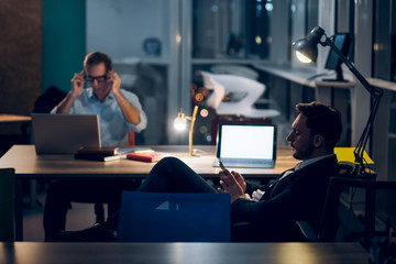 Two tired colleagues working late night. Handsome young businessman with beard typing message on his phone while sitting at office desk with his coworker across the table in background.