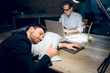 © Svyatoslav Lypynskyy - Young man fell asleep in office at night. Good looking businessman resting his head on pillow on brown wooden office desk. His coworker sitting with laptop working in background.