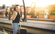© terovesalainen - Woman waiting for train in station. Lady standing in platform with suitcase, passport and ticket. Happy female traveler going on vacation at sunset.
