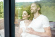 Young and lovely couple standing together looking outside the window at home, view through the window vith reflection