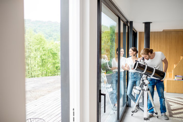 Young couple watching with telescope standing near the window in the modern house