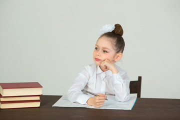 Wall Mural - beautiful little girl with a bow on her head sitting at a desk with books on a white background and doing homework