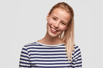 Wall Mural - Happy female student with broad smile, has light hair combed in pony tail, recieves positive news, dressed in striped casual jacket, glad to meet with best friend, isolated on white background