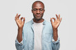 Portrait of pleased attractive African American guy makes okay gesture, has dark skin, smiles gently, eyes closed, wears denim shirt, isolated over white background. People, body language concept