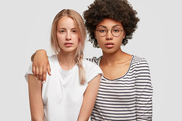 Wall Mural - Attractive mixed race women embrace and have good relationship, being best friends, stand together against white wall. Pretty multiethnic lesbians spend free time in friendly company. Friendship