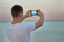 Guy Tourist In A White Shirt Takes Pictures Using A Smartphone Stone In The Sea At Sunset. The Dead Sea, Israel. Dreamy Mountain Landscape Of Jordan, Rear View. Concept Technology And Smartphones