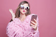 Gorgeous lady dressed in funky bushy coat, wears make up, makes video call via smart phone and headphones, looking at gadget screen, taking selfie sending kiss to boyfriend, standing over pink wall..