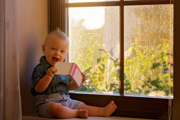 Canvas Print - Baby boy, cute child, sitting on a window on sunset, playing with baby book