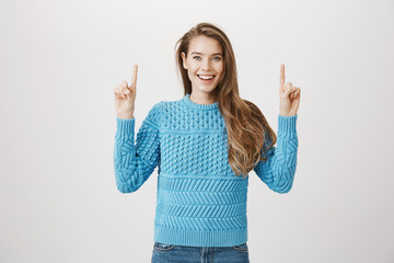 Portrait of positive adorable blonde model pointing up while being excited and happy with what she sees, smiling broadly while standing against gray background. Look there, you would not regret