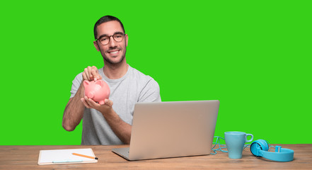 Happy young man sitting at his desk and holding a piggy bank - Green background