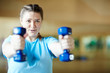 © pressmaster - Young female in blue t-shirt doing effective exercise with dumbbells while working out in gym