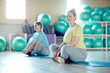 Young chubby cross-legged woman and her groupmate in activewear sitting on mats while exercising in fitness center