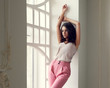 © Dmitry Tsvetkov - Young stylish beautiful woman wearing pink trousers and silk blouse. Casual style. Pretty tanned girl with wavy brunette hair standing and posing near white window and relaxing