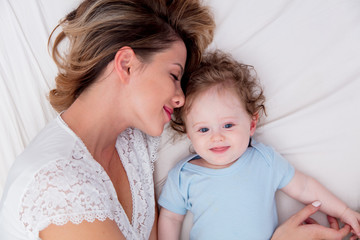 Wall Mural - Young mother with her 7 month old little son dressed in pajamas are relaxing and playing in the bed at the weekend together, lazy morning, warm and cozy scene.