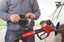 Lack Of Money For Gasoline And Fuel. Expensive Gasoline. Driver Man Holds One Dollar End Empty Wallet Against The Background Of A Fuel Nozzle In The Gas Tank. Increase In Gasoline Prices Concept.