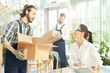 © pressmaster - Smiling pretty young female entrepreneur in glasses interacting with movers and getting goods, she making notes in paper while viewing goods in lobby