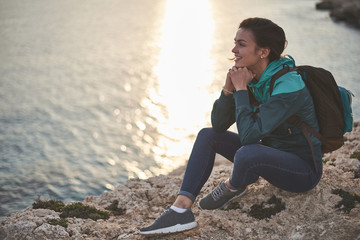 Wall Mural - Traveling brings pleasure. Cheerful young woman is enjoying sunset near the ocean. She is sitting on stony hill and smiling 