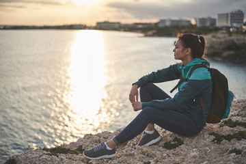 Wall Mural - Calm girl is watching sunset at the seashore. She is sitting on stone with beautiful landscape on background 